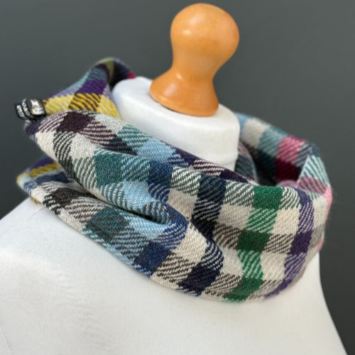 Multicolored plaid scarf on a mannequin with a wooden top against a gray background. The scarf features blue, green, and purple hues.