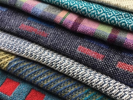 Stack of folded fabrics with colourful patterns: stripes, checks, and geometric designs. Dominant hues include blue, red, and green.