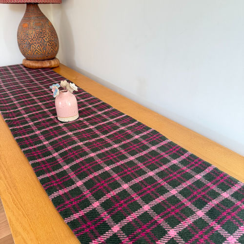 Wooden table with a black-and-pink plaid runner, a small pink vase of flowers, and a patterned lamp base against a plain wall.