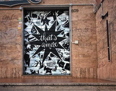 Black and white street art on a metal shutter in Rome, featuring repeating Günter skulls and eyes with the phrase "That's Amore" over a starburst.