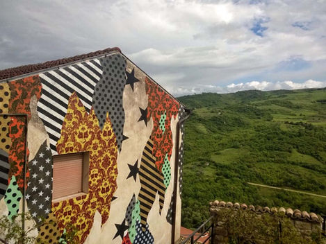 Veduta grandangolare del murale 'CAMOUFLAGE 86030' su una casa a Civitacampomarano, che contrasta con i motivi vivaci rispetto al paesaggio urbano tradizionale.