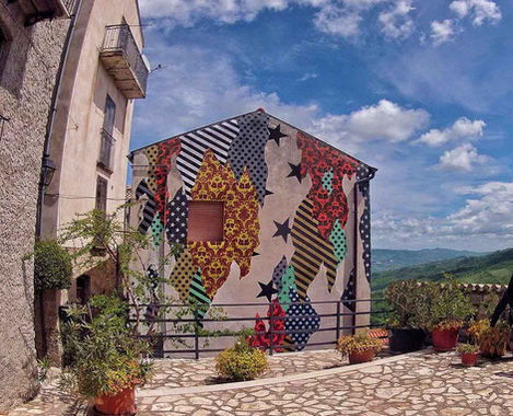 Veduta completa del murale 'CAMOUFLAGE 86030' di UNO su facciata bianca, che mostra sezioni a righe e pattern vari, con vista sulle colline verdi.
