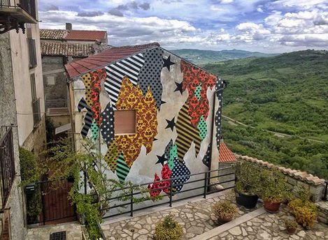 Veduta grandangolare del murale 'CAMOUFLAGE 86030' su casa a Civitacampomarano, che contrappone pattern vivaci al paesaggio urbano tradizionale.