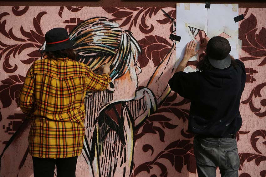 Dettaglio murales: ragazza che disegna di Alice Pasquini su sfondo damascato pixel di UNO, Roma.