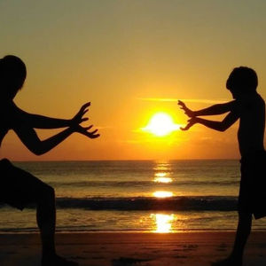 Silhouettes of two boys facing each other and holding their hands out towards one another with the rising sun and the ocean in the background. 
