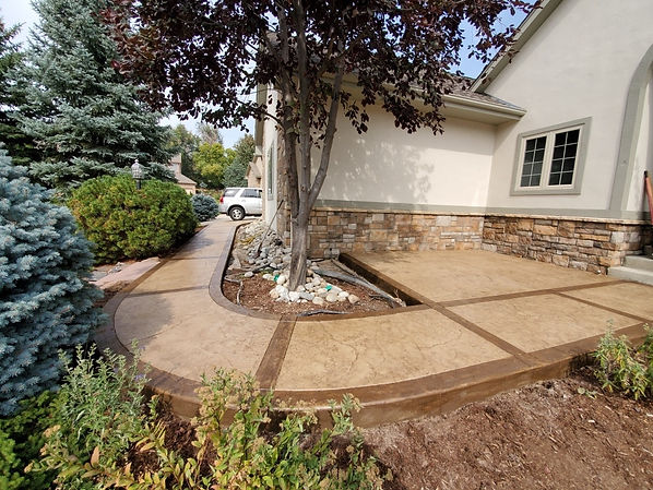 A two-tone brown and light brown stamped concrete walkway poured around existing landscaping at a Colorado home.