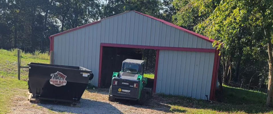 A red-trimmed gray shed with a green utility vehicle parked inside. A black dumpster with a "Clean Slate" logo is nearby, on a grassy lot.