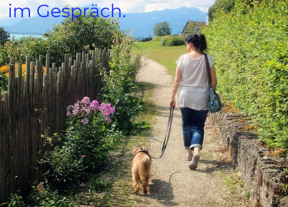 Frau lÀuft mit kleinem Hund an der Leine einen befestigten Weg in gepflegter Natur vor einem Berghintergrund.