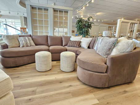 A large, curved taupe sectional sofa arranged with two round cream ottomans on a light wood floor inside the Werner Harmsen furniture showroom in Waupun.