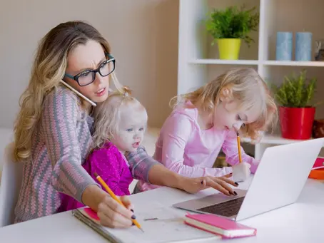 Busy mom multitasking at home while working on a laptop, caring for two young children, and writing notes—illustrating the challenges of balancing work and family life.