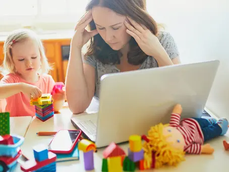 Busy mom multitasking at home while working on a laptop, caring for two young children, and writing notes—illustrating the challenges of balancing work and family life.