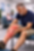 Man in blue shirt holds his red, swollen leg in a waiting room. Background shows seated couple, plants, and posters.