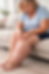 Elderly woman in blue shirt sits on beige couch, massaging her swollen leg. Living room setting with a plant in the background.