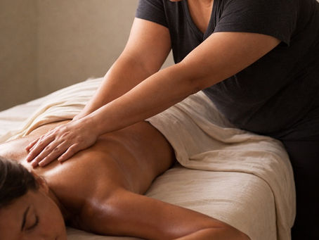 A massage therapist in a dark shirt gives a relaxing back massage to a client on a white massage table in a calm, neutral-toned room.