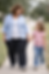Woman and girl smiling, holding hands while walking on a tree-lined sidewalk. The woman wears a blue shirt, the girl a pink striped tee.