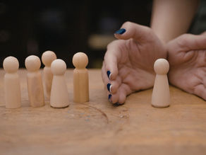 Wooden figures on a table; one separated by hands with dark blue nails. Brown background, creating a mood of distinction or separation.