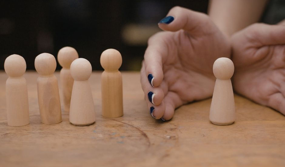 Wooden figures on a table; one separated by hands with dark blue nails. Brown background, creating a mood of distinction or separation.