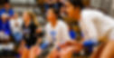 Three female volleyball players in a gym crouch in ready positions, focused on the game, with spectators seated on bleachers in the background.