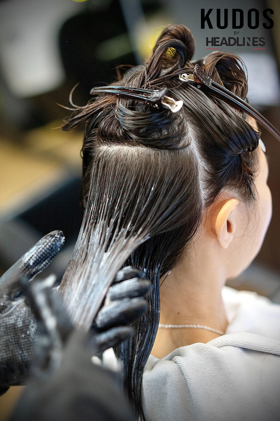 Hair treatment in progress: person with clipped hair, stylist applying product with gloved hands. Logo "KUDOS by HEADLINES" visible.