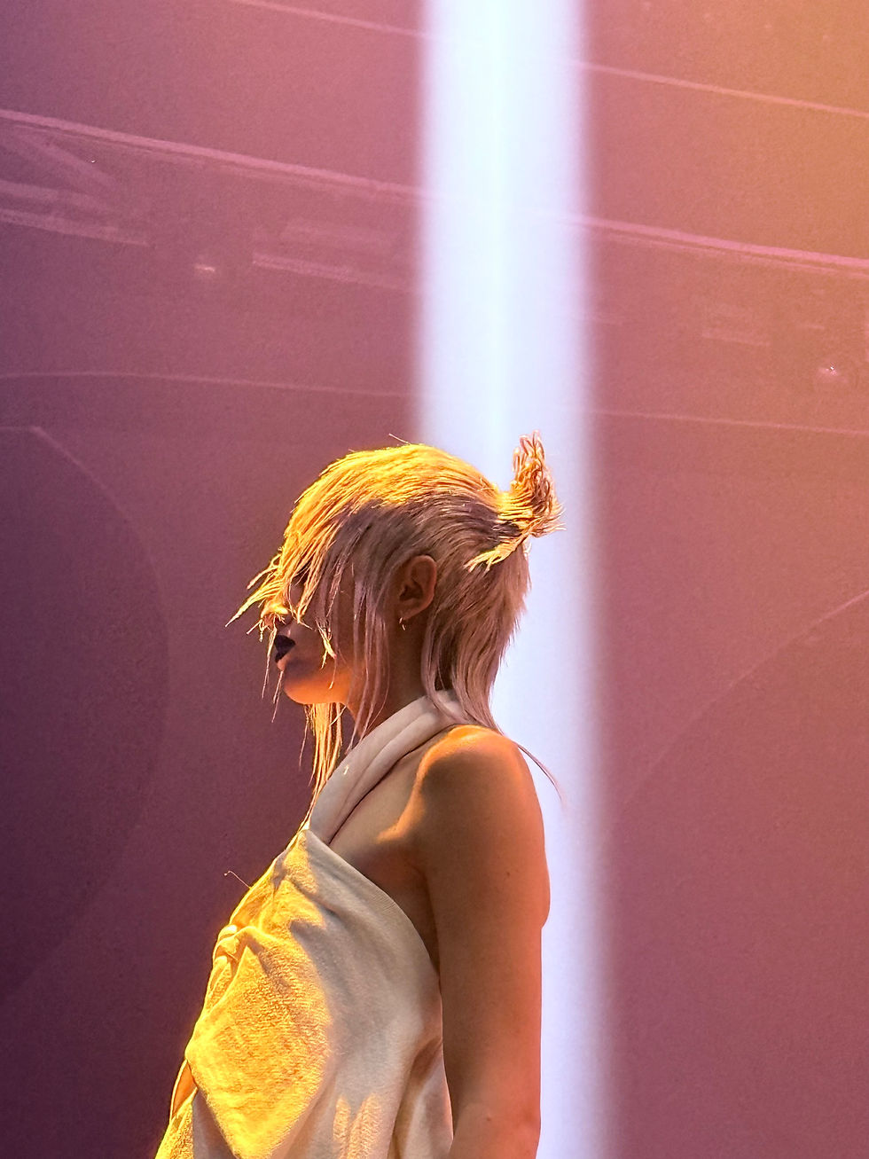 Person with platinum hair in a white draped outfit stands in a warm spotlight against a purple-pink stage backdrop.