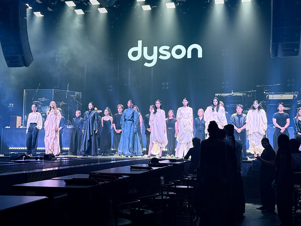 Models stand on a lit runway stage beneath a large Dyson logo, with audience silhouettes in front and blue haze.
