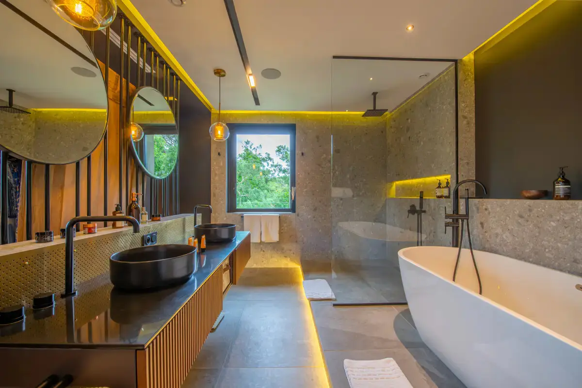 Salle de bains chambre de maître avec baignoire ovale qui repose au sol. Deux vasques lavoabo de forme arrondie reposent sur un plan horizontal dans les tons de brun. Douche à l'italienne.