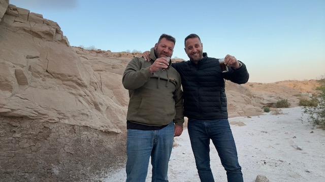 Amir and Arik raising a toast in a sandy wash at dusk