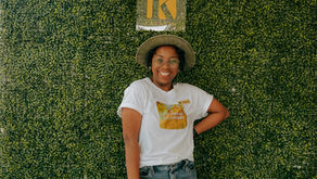 Mel Jeanty in a hat, wearing a white shirt and jeans, poses against a green leafy wall at Kayasah in Haiti. The yellow Kayasah logo is visible above.