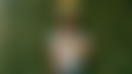 Mel Jeanty in a hat, wearing a white shirt and jeans, poses against a green leafy wall at Kayasah in Haiti. The yellow Kayasah logo is visible above.