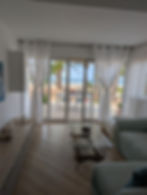 Bright living room with white curtains, glass table, and a sofa. Patio doors open to a beach view with palm trees, sand, and ocean.