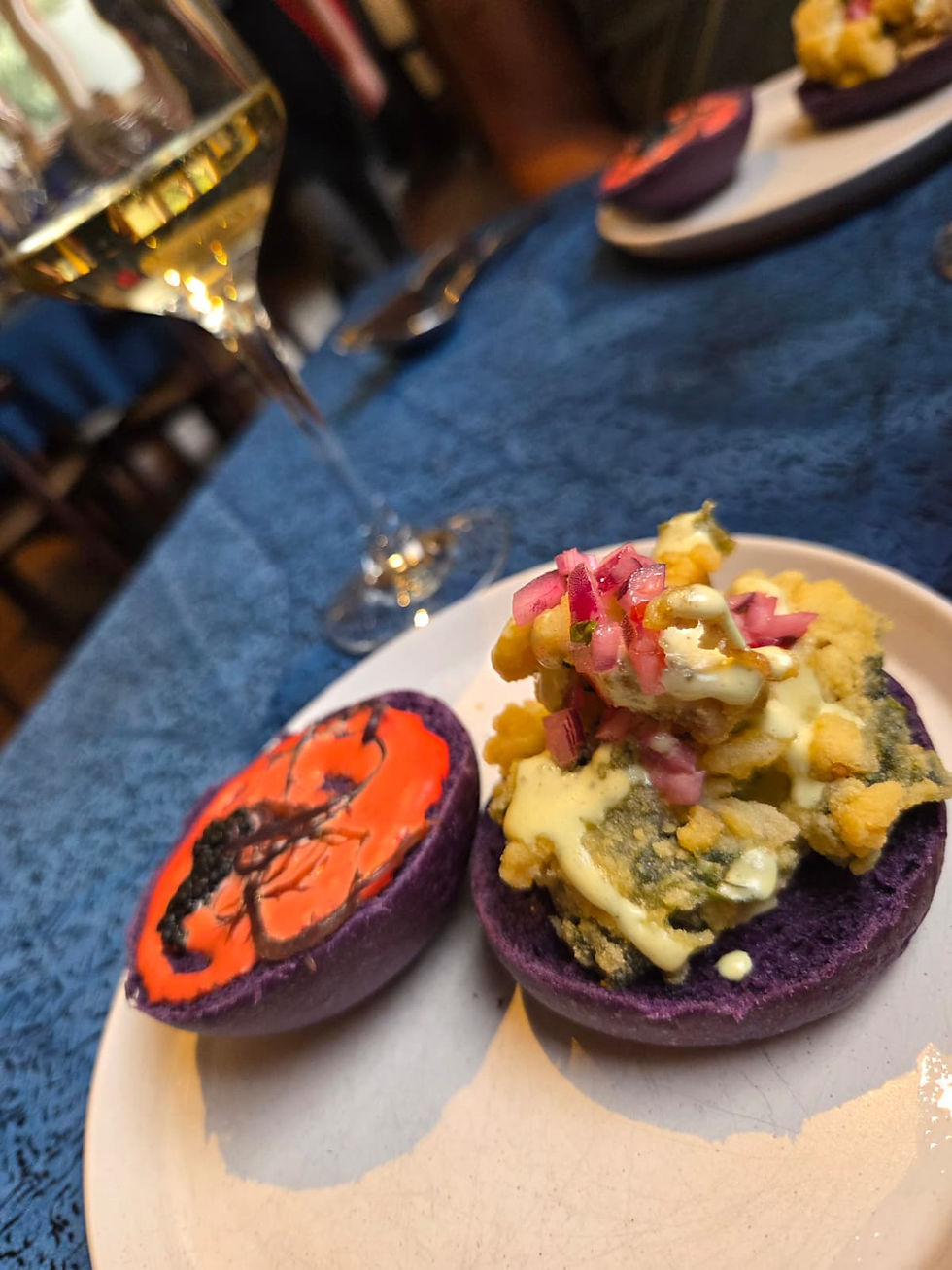 Purple bao buns topped with fried seafood, pickled onions and sauce on a white plate beside a wine glass.
