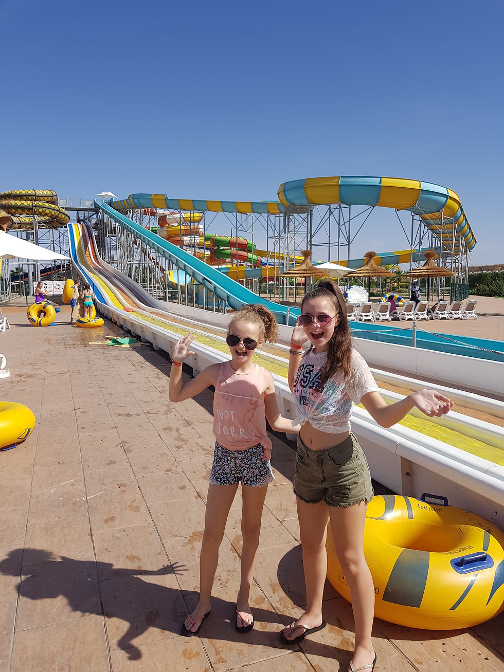 Two girls pose happily by a colorful water slide at a sunny water park, wearing casual summer outfits. Bright yellow tube and slides in view.