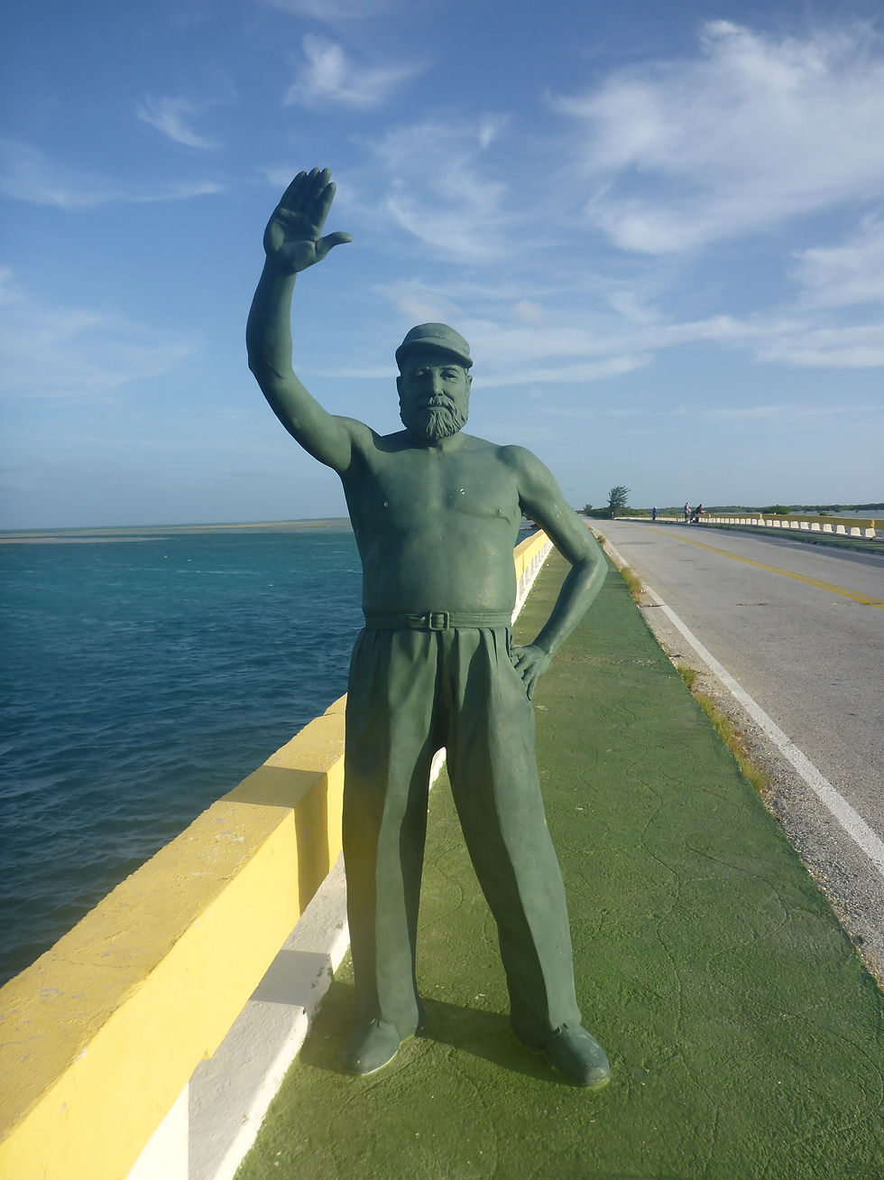 Statue of a man waving stands on a green path by the ocean. Bright blue sky, yellow railing, and distant figures on a road.