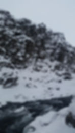 Snow-covered rocky cliff with a dark, flowing river below. Overcast sky creates a tranquil, cold atmosphere.