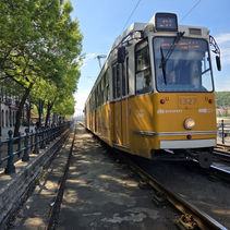 Yellow Budapest tram 1327 on riverside tracks under trees on a sunny day, with overhead wires and a driver visible.