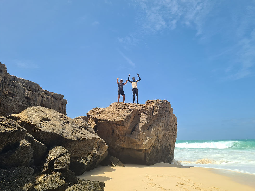 Two people stand on a rocky cliff, arms raised, overlooking a sunny beach with waves. Clear sky and sandy shoreline in the background. Boa Vista, Cape Verde