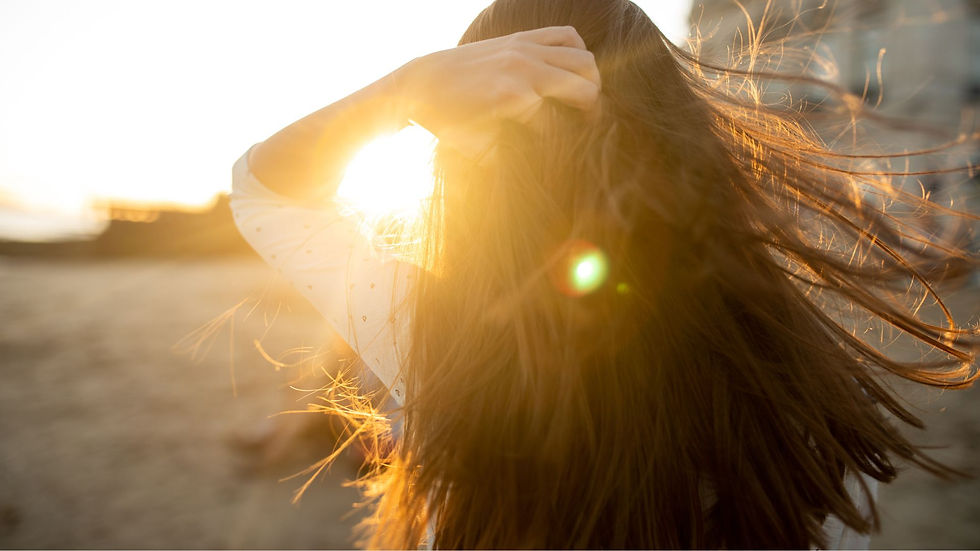 A woman standing on a beach with sunlight coming through her hair that is being wind blown.