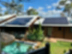 Eco-friendly wooden house with solar panels, surrounded by trees. Features a pool and a thatched area in sunny backyard. Relaxed setting.
