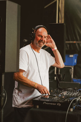 A bald man with headphones on stands behind a DJ set and speakers.