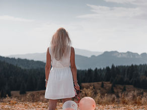 A woman in a flowing white dress stands on a rocky hilltop, gazing at the distant mountains and sky. She holds a small cluster of peach and white balloons, symbolizing dreams, aspirations, and new beginnings. The peaceful landscape and soft lighting evoke a sense of reflection and possibility—perfect for embracing big dreams and setting meaningful life goals for the year ahead.
