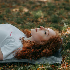 A woman with curly red hair lies on a blanket in soft afternoon light, eyes closed and breathing deeply, resting on the grass beneath scattered  leaves — a peaceful moment of exhaustion giving way to calm.
