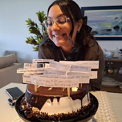 Letícia é parda, de cabelo preso pela metade, castanho. Ela está na frente de um bolo com placas de tags do AO3, sorrindo.