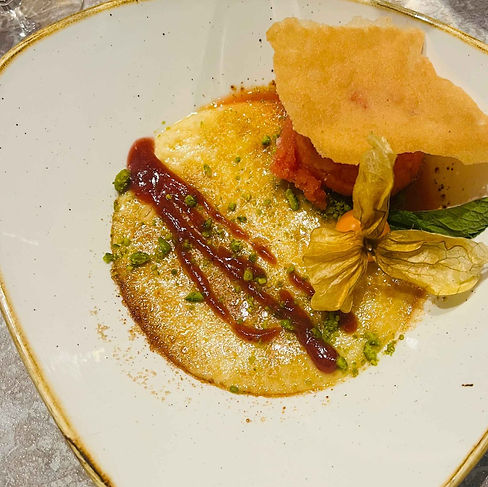 Fein angerichtetes Dessert auf einem cremefarbenen Teller mit karamellisierter Oberfläche, Fruchtsauce, Pistazienstückchen und essbarer Blüte als Dekoration.