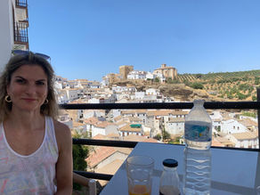 Jordana smiling at a café table overlooking Setenil de las Bodegas, a white hillside town in southern Spain, with an old castle and church visible on the rocky outcrop, orange juice and water bottle on the table.