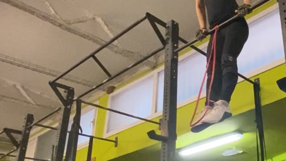 Woman performing a bar muscle-up above the bar at a CrossFit box in Barcelona, resistance band visible but not load-bearing