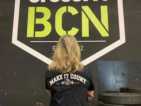 Woman standing in front of the CrossFit BCN logo wall wearing a Make It Count t-shirt, surrounded by weight plates.