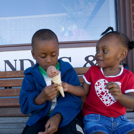 Two young children sitting on a wooden bench. A young girl smiles as she offers her chocolate ice cream code to a little boy.