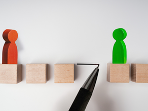 A conceptual image showing two wooden figurines, one red and one green, standing on separate wooden blocks with a gap between them. A pen is drawing a bridge between the blocks, symbolizing connection, communication, or bridging understanding between two perspectives.