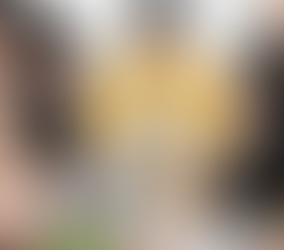 A group of diverse professionals are gathered around a table in a collaborative workspace. At the center, a Black woman wearing a bright yellow cardigan stands with a serious, thoughtful expression, leaning forward with her hands on the table. She appears to be leading the discussion or considering a complex point as the others look at her or down at laptops and documents. The group includes men and women of various racial backgrounds, dressed casually, suggesting an informal yet focused team meeting. The table is cluttered with color swatches, notebooks, coffee cups, and laptops, indicating a creative or strategic work session. The mood is intense and reflective, capturing the emotional weight of decision-making and leadership in a diverse team setting.