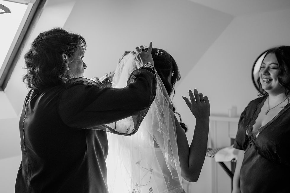 Black-and-white photo of wedding prep as the bride's mother adjusts her veil while her bridesmaid watches in a bright room.
